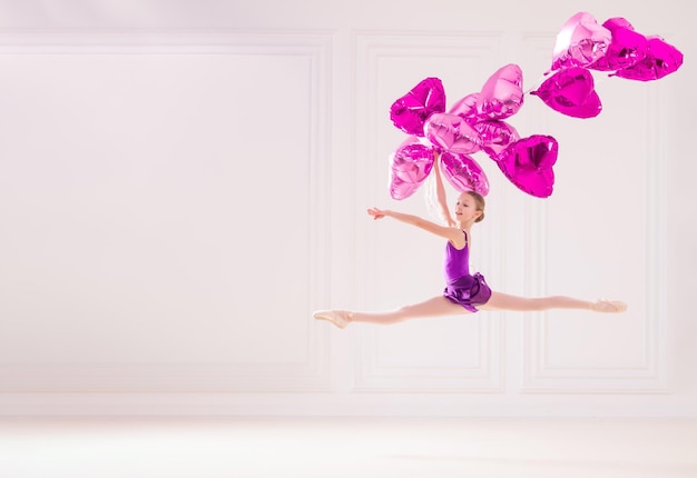 Estudiante de bailarina en traje de baño lila vuela con un montón de globos rosas en forma de corazón en una hermosa habitación blanca