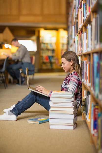 Estudiante bastante feliz que usa la computadora portátil en el piso de la biblioteca