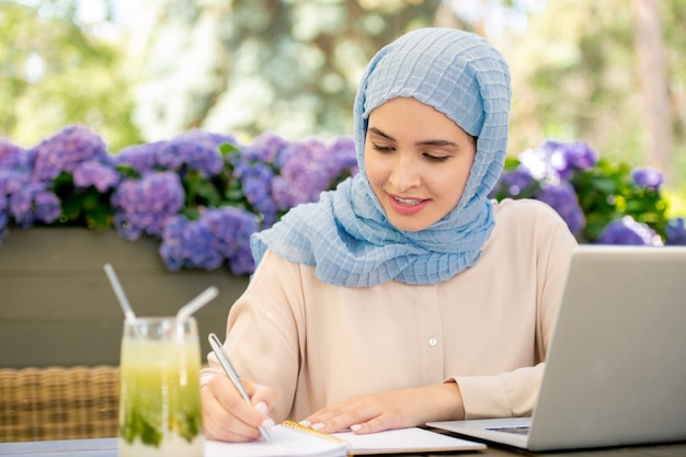 Estudiante bastante musulmán en hijab tomando notas en el cuaderno mientras está sentado frente a la computadora portátil mientras trabaja en un proyecto al aire libre