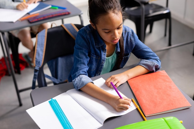 Foto una estudiante bi-racial feliz sentada en el escritorio y tomando notas en el aula de la escuela