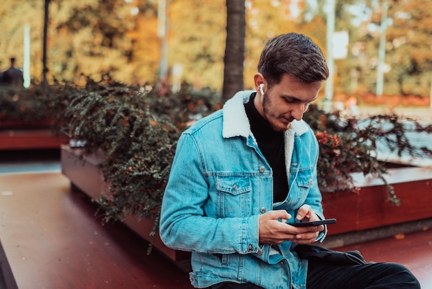 Un estudiante boquiabierto sentado en un parque usando un teléfono inteligente y auriculares inalámbricos
