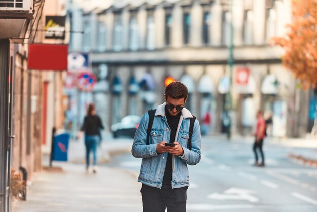 Un estudiante caminando por la ciudad con una mochila y gafas de sol mientras usa un smartpho