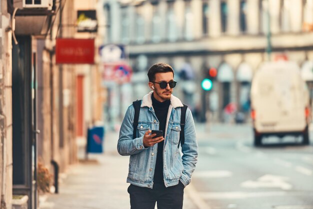 Un estudiante caminando por la ciudad con una mochila y gafas de sol mientras usa un smartpho