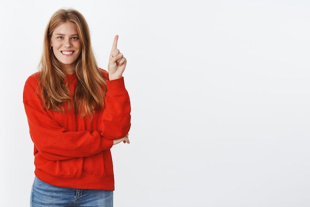 Estudiante carismática y ambiciosa con lindas pecas, ojos azules profundos y cabello rojo natural, de pie en un acogedor suéter rojo, apuntando hacia arriba y sonriendo ampliamente, dando consejos para usar un espacio de copia increíble