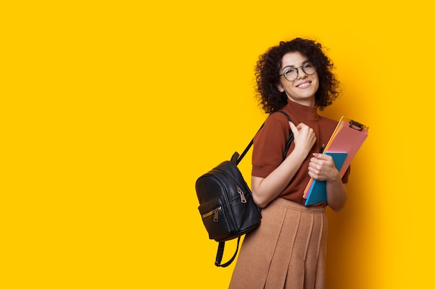 Estudiante caucásico alegre con una bolsa y algunos libros está posando feliz sobre un fondo amarillo con espacio libre