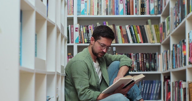 Estudiante caucásico hombre leyendo un libro sentado entre una pila de libros en la vista lateral del piso de la biblioteca