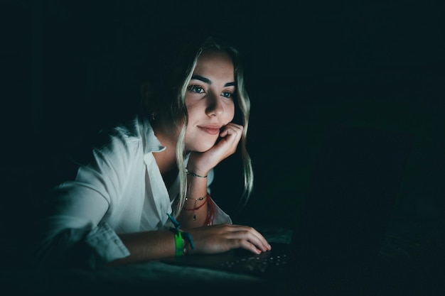 Estudiante caucásico sonriente y confiado que usa una computadora portátil por la noche en la cama trabajando terminando un proyecto a altas horas fecha límite mirando la pantalla de la computadora sentado en el dormitorio para la tarea universitaria