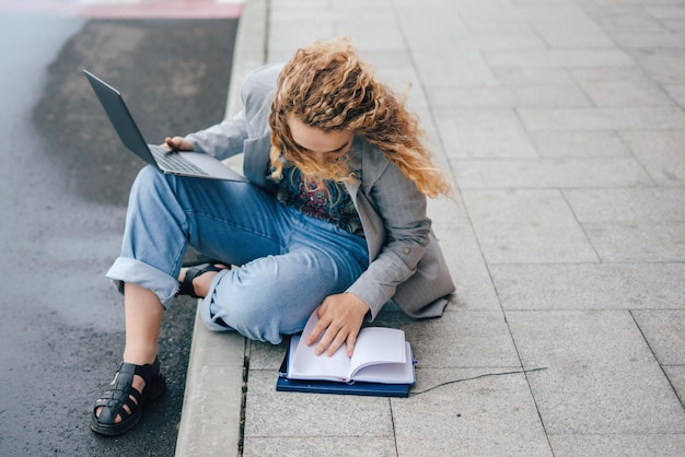 Estudiante con chaqueta estudiando con laptop y bloc de notas sentada al aire libre en la calle de la ciudad preparándose para los exámenes Estudiar desde casa para prevenir la infección por el virus Corona o el brote de Covid19