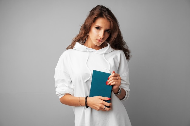 Estudiante chica adolescente con capucha blanca sosteniendo un libro en sus manos sobre un fondo gris