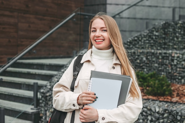 Estudiante chica feliz sosteniendo carpetas de cuaderno en las manos, sonriendo en el edificio de la universidad