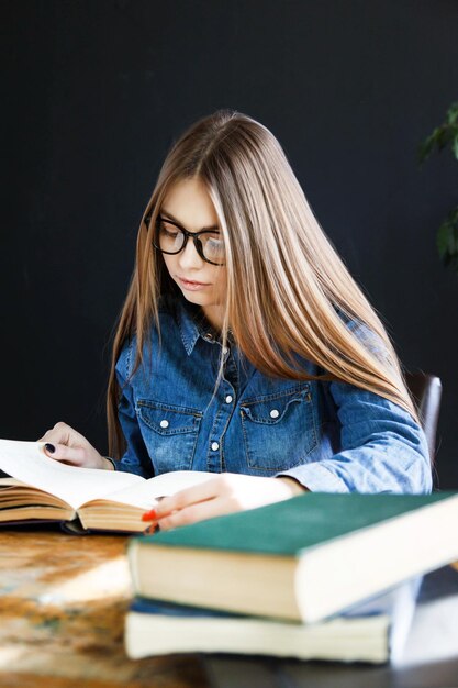 Estudiante chica de pelo largo con gafas sentada junto a una mesa de madera y leyendo un libro