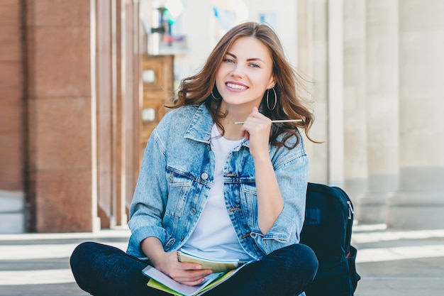 Estudiante chica sentada cerca de la universidad y sonriendo.