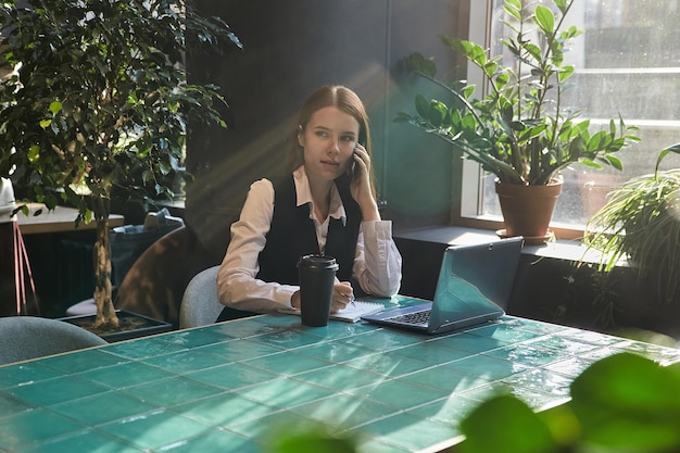 Estudiante chica sentada en la mesa de café estudiando con laptop llamando por teléfono inteligente