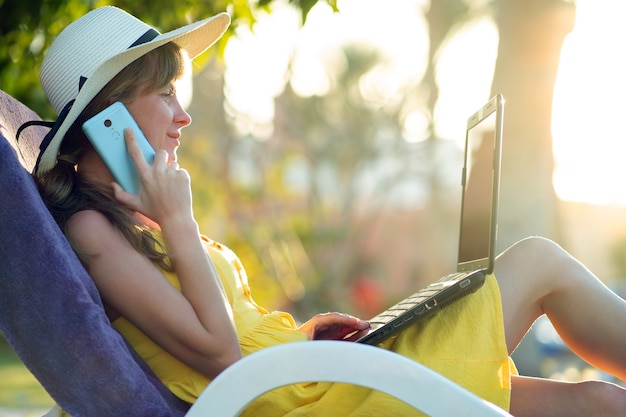 Estudiante chica en vestido amarillo de verano descansando sobre césped verde en el parque de verano estudiando en la computadora portátil que tiene una conversación en el teléfono celular móvil.