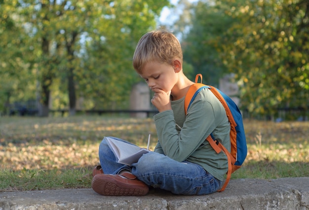 Estudiante chico europeo haciendo los deberes en un cuaderno, sentado en un hermoso parque otoñal