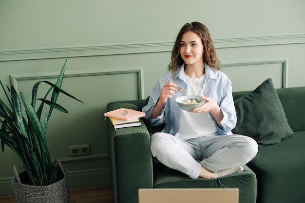 Estudiante contenta tomando un descanso de las clases en línea, libros de comida y una computadora portátil en el sofá, mujer feliz