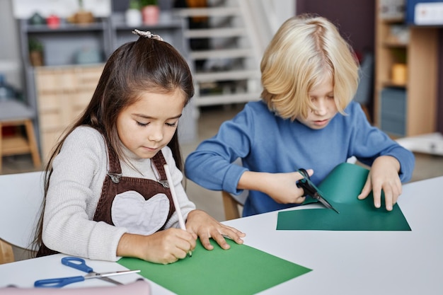 Estudiante diligente de la escuela primaria dibujando en papel verde oscuro mientras crea juguetes con clase rubia