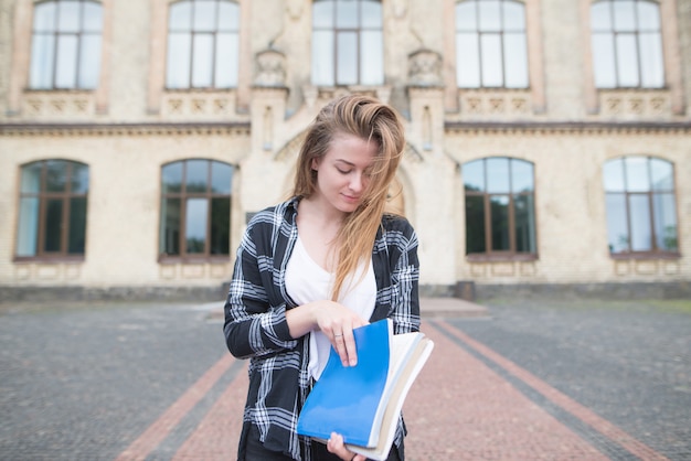 Estudiante en un elegante vestido casual con libros y cuadernos en sus manos contra el fondo de un edificio universitario.