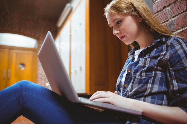 Estudiante enfocado que se sienta en el piso contra la pared usando la computadora portátil