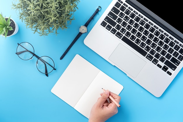 Un estudiante escribe en un libro blanco abierto o contabilidad aislada en un lugar de trabajo azul limpio mínimo en casa con teléfono inteligente y accesorios, espacio de copia, endecha plana, vista superior,