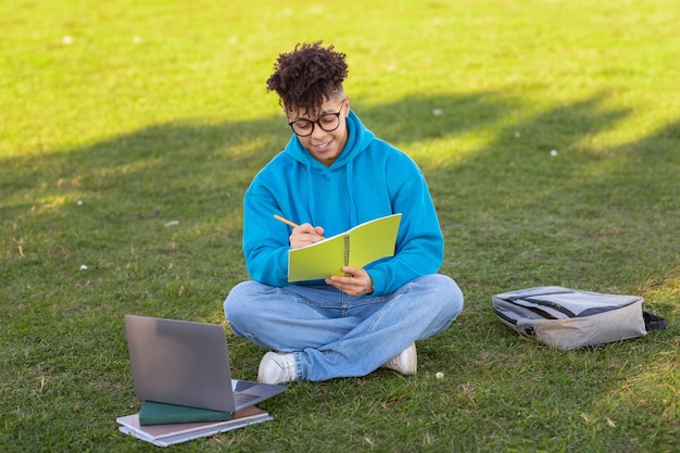 Estudiante escribiendo en un cuaderno con una computadora portátil al lado