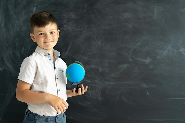 Estudiante de escuela primaria de pie con un globo terráqueo cerca de una pizarra en un salón de clases con una pizarra en el fondo Lección de geografía para escolares Niño en la universidad
