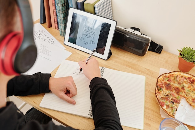 Estudiante de la escuela sentado en su escritorio en casa, viendo el curso de programación en línea y el esquema de dibujo en el libro de texto