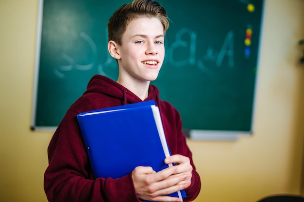 Estudiante de la escuela superior está de pie en una sudadera con capucha oscura cerca de la pizarra verde en el aula Adolescente con cuaderno azul en las manos está mirando a la cámara Primer plano