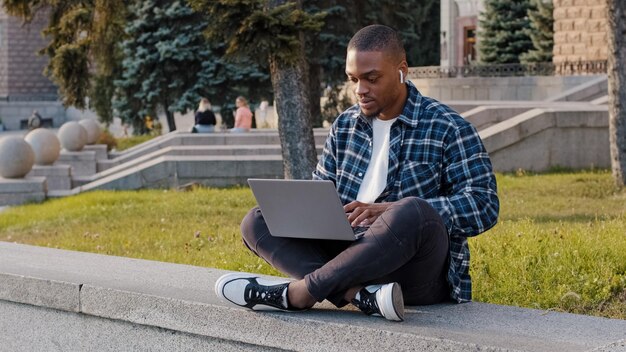 Estudiante estadounidense chico hombre de negocios africano sentado al aire libre en la calle de la ciudad escribiendo en la computadora portátil trabajando