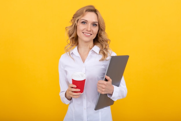 Una estudiante feliz con una laptop sostiene una bebida de café para llevar en un vaso de papel con fondo amarillo