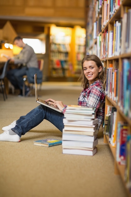 Foto estudiante feliz que usa la computadora portátil en el piso de la biblioteca que mira la computadora portátil