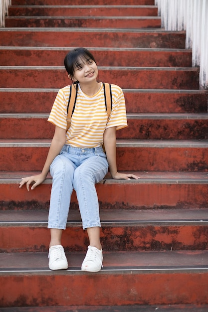 Estudiante feliz usa camisa amarilla y jean con mochila sentado en las escaleras de la escuela.