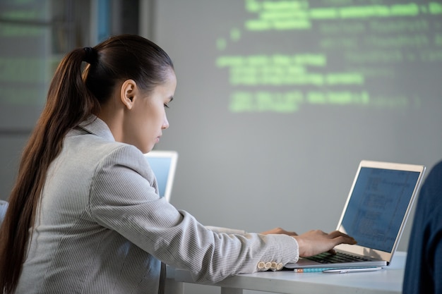 Estudiante femenina contemporánea de la universidad técnica sentada frente a la computadora portátil y la pantalla mientras trabaja sobre la presentación del nuevo software