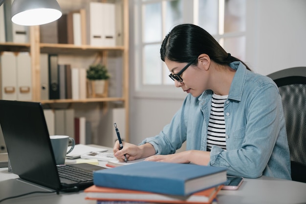 Estudiante femenina tomando notas del libro en casa. Joven japonesa asiática sentada en la mesa haciendo tareas para el proyecto de examen final en la escuela. vista lateral de una elegante y elegante colegiala haciendo los deberes.