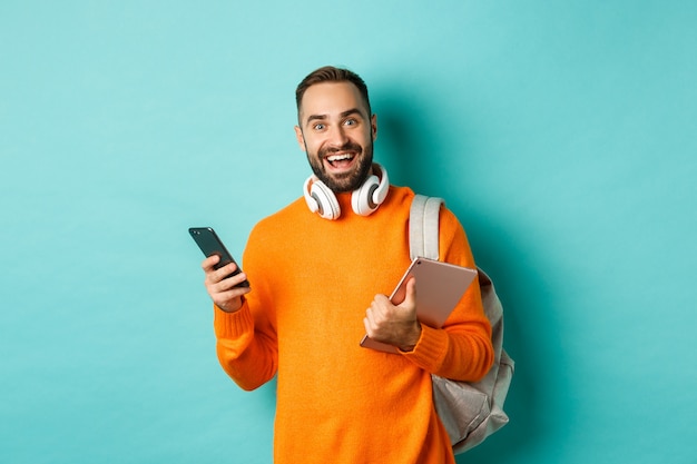 Estudiante guapo con auriculares y mochila, sosteniendo tableta digital y teléfono inteligente, mirando a cámara, de pie contra el fondo turquesa.