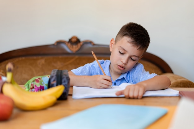 Foto el estudiante hace su tarea, escribe con cuidado en un cuaderno grande, trabaja en casa en una gran mesa de madera y tiene plátanos al lado de sus cuadernos por si tiene hambre.