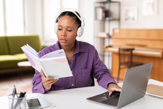 Una estudiante haciendo varias tareas con un libro y una computadora portátil.