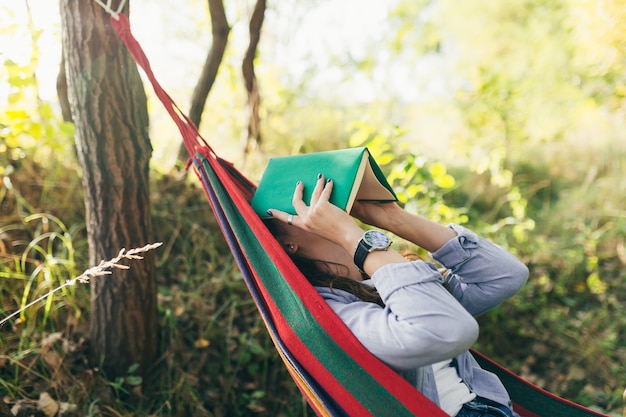 Estudiante hermosa chica descansando en el parque tumbado en una hamaca y leyendo un libro