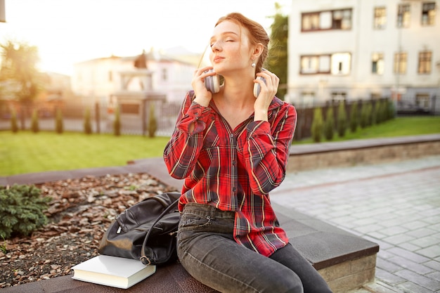 Foto estudiante hermoso joven que lleva auriculares de una camisa a cuadros y una mochila que descansa sobre la calle