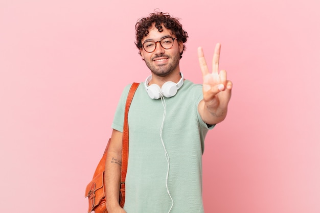 Estudiante hispano sonriendo y luciendo feliz, despreocupado y positivo, gesticulando victoria o paz con una mano