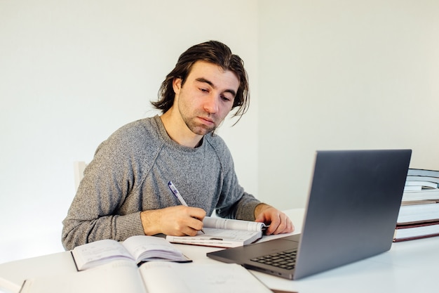 Estudiante hombre escribe en el cuaderno trabajando en equipo. Marketing en Internet, trabajo independiente, trabajo desde casa, aprendizaje en línea, estudio, concepto de bloqueo. Educación a distancia, libros sobre la mesa