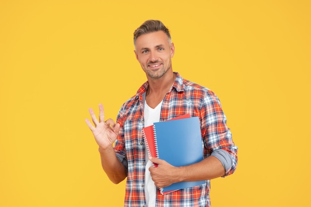 Estudiante hombre feliz mostrando gesto de anillo OK sosteniendo libros universidad de fondo amarillo