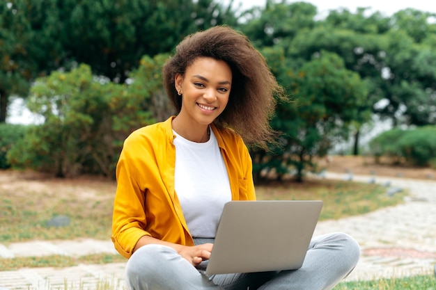 Una estudiante independiente afroamericana de cabello rizado, de moda, positiva, con ropa informal y elegante, sentada con una laptop al aire libre mientras trabaja o estudia en línea, mira las sonrisas de la cámara