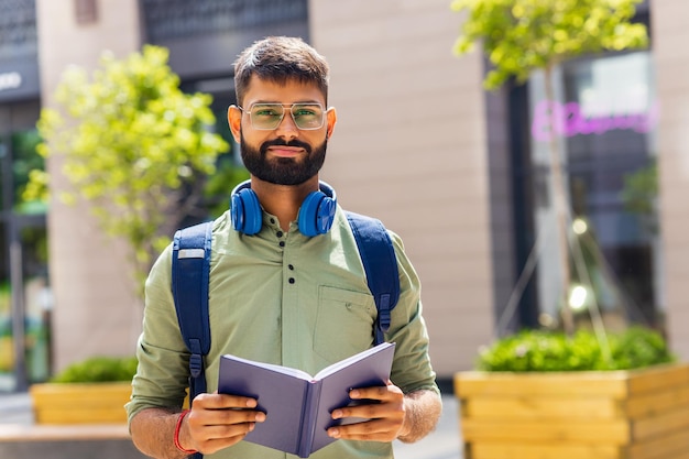 Estudiante indio con auriculares azules y mochila con libros en un día soleado