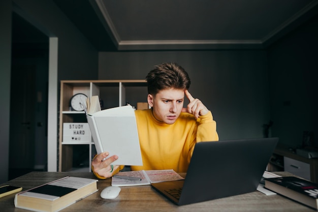 Estudiante inteligente sentado en casa en la mesa en el fondo de la habitación con un libro en la mano y mirando la pantalla de la computadora portátil con una cara seria Un tipo pensativo estudia de forma remota en Internet