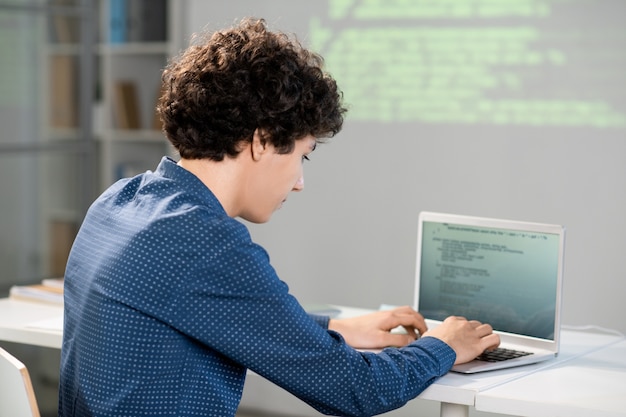 Estudiante inteligente serio inclinándose por el escritorio frente a la computadora portátil mientras prepara la presentación de su proyecto para la conferencia después de clases