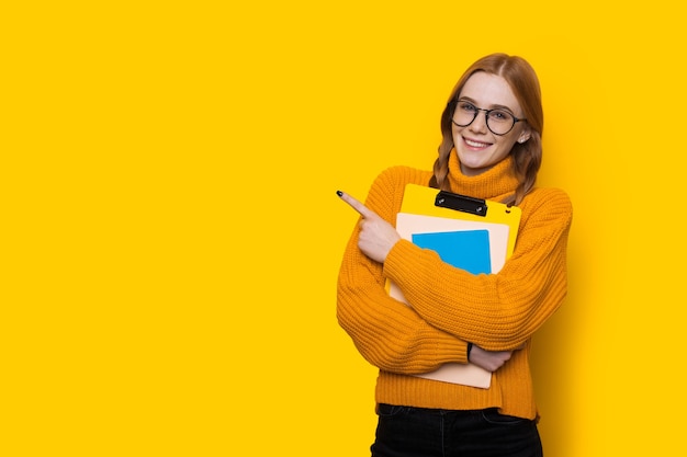 Estudiante de jengibre con pecas y anteojos apuntando hacia el espacio libre amarillo cerca de ella con el dedo abrazando algunos libros