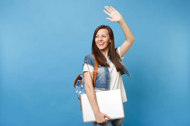 Estudiante joven alegre feliz con mochila agitando la mano para saludar, conocer amigos con ordenador portátil aislado sobre fondo azul. Educación en la universidad. Copie el espacio para publicidad.