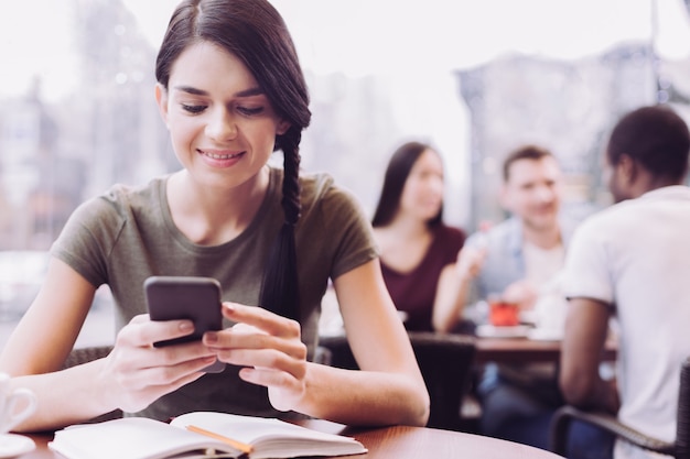 Estudiante joven alegre que usa el teléfono mientras hace la tarea y mira hacia abajo