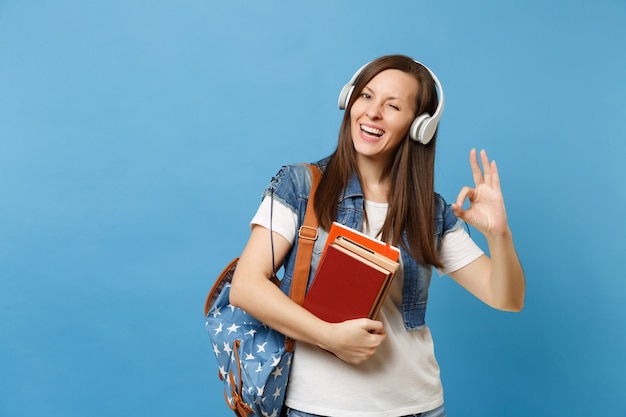 Estudiante joven alegre en ropa de mezclilla con auriculares mochila escuchar música mantenga libros escolares mostrando signo OK parpadeando aislado sobre fondo azul. Educación en el colegio universitario de secundaria.